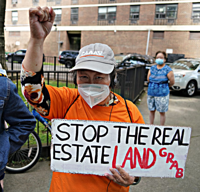 Member of CAAAV holding a sign that says STOP THE REAL ESTATE LAND GRAB, wearing CAAAV hat and shirt, raising right fist
